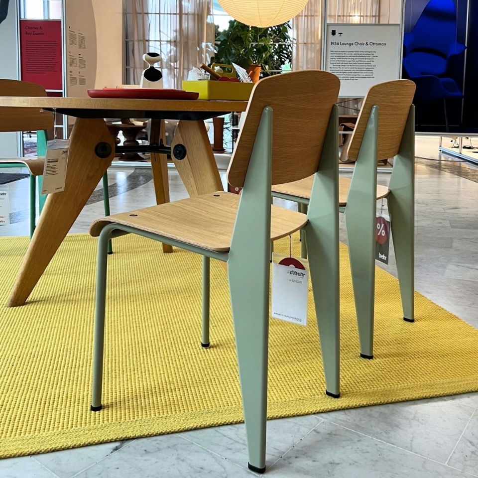 Set of light wood chairs with mint-green legs arranged around a wooden table in a showroom, price tags hanging from the chairs on a yellow rug.