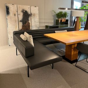 Modern black leather sectional sofa with metal legs around a large wooden dining table in a showroom, with cushions and abstract art panels in the background.