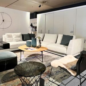 Modern showroom living room with white sectional sofa, teal vases on a round wooden coffee table, and a geometric wire chair nearby.