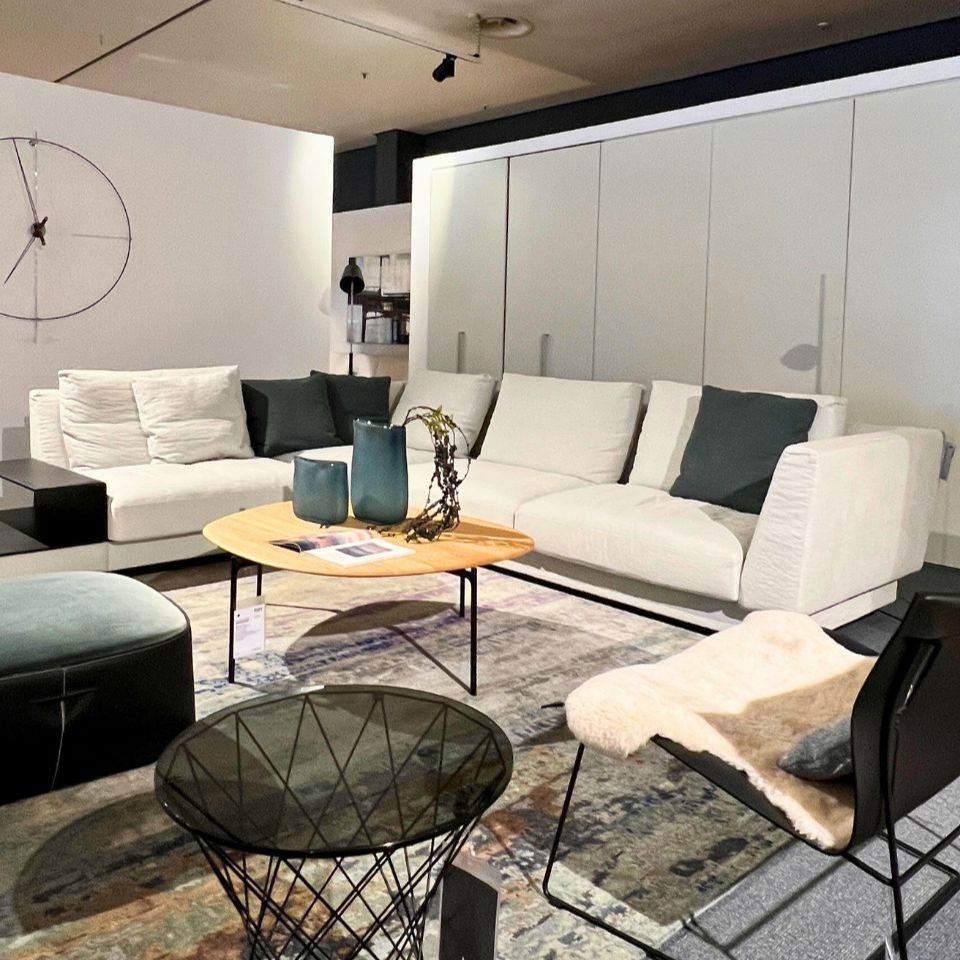 Modern showroom living room with white sectional sofa, teal vases on a round wooden coffee table, and a geometric wire chair nearby.
