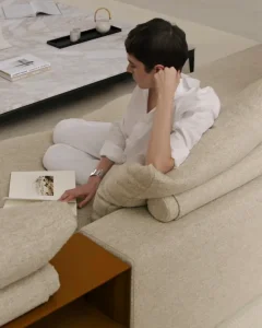 Person in white clothing sits on a cream sofa, reading an open book with one hand resting on their head.
