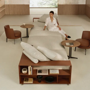 Person in a white robe sits on a striped bed in a bright, minimalist lounge with round wooden tables and brown chairs nearby.