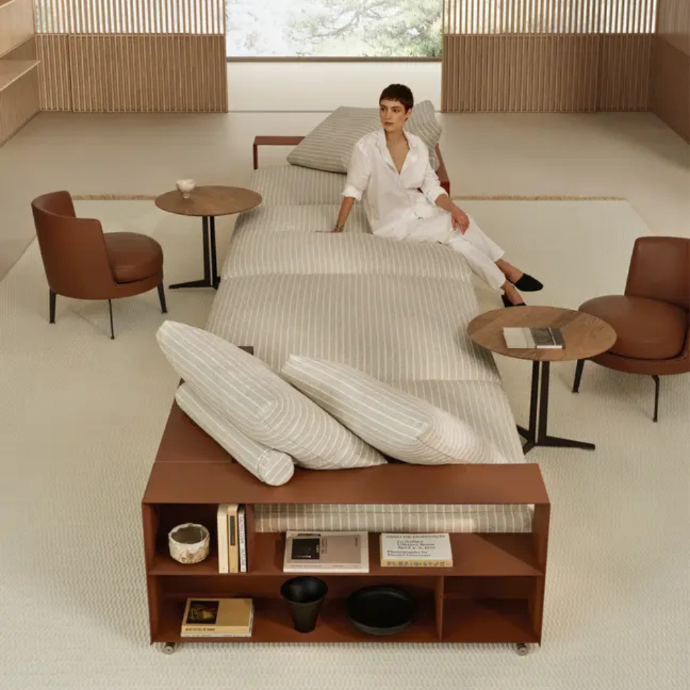 Person in a white robe sits on a striped bed in a bright, minimalist lounge with round wooden tables and brown chairs nearby.