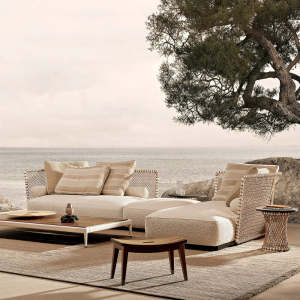 Coastal outdoor lounge with beige cushioned sofa, rattan chairs, and a wooden coffee table overlooking the sea under a large tree branch.