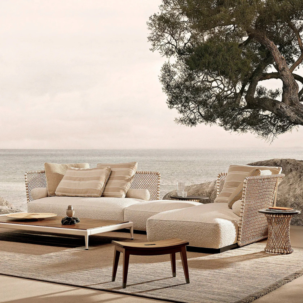 Coastal outdoor lounge with beige cushioned sofa, rattan chairs, and a wooden coffee table overlooking the sea under a large tree branch.