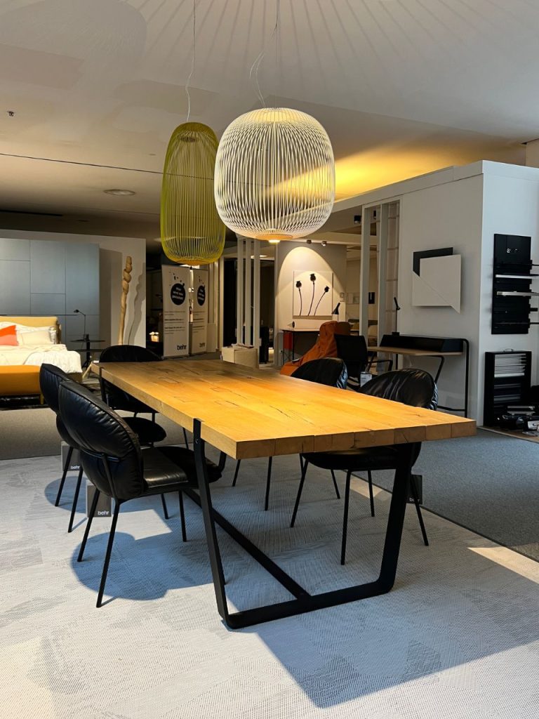 Modern showroom with a long wooden dining table and black upholstered chairs beneath sculptural pendant lights in green and white shapes.