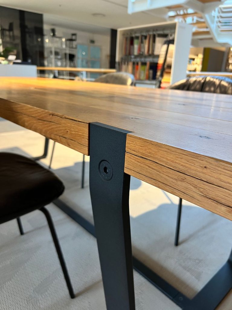 Close-up of a wooden table edge with a dark metal leg and bolt in a modern communal workspace with shelves in the background.