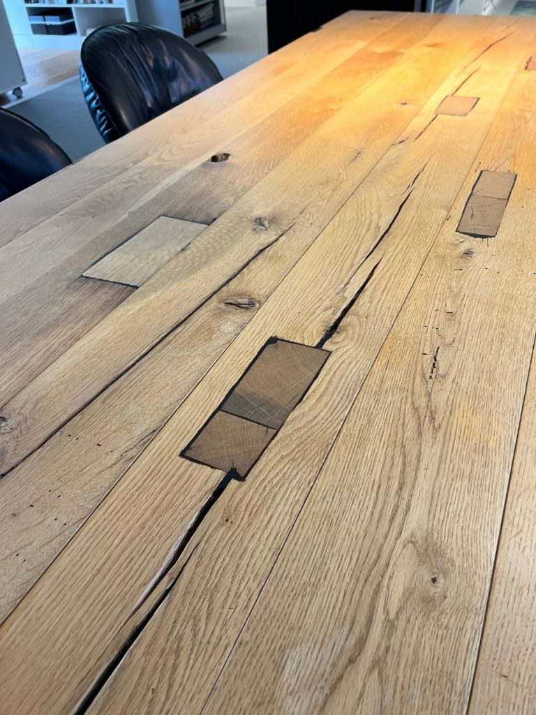 Long wooden conference table with visible cracks and patched-in boards, surrounded by black leather chairs in an office setting.