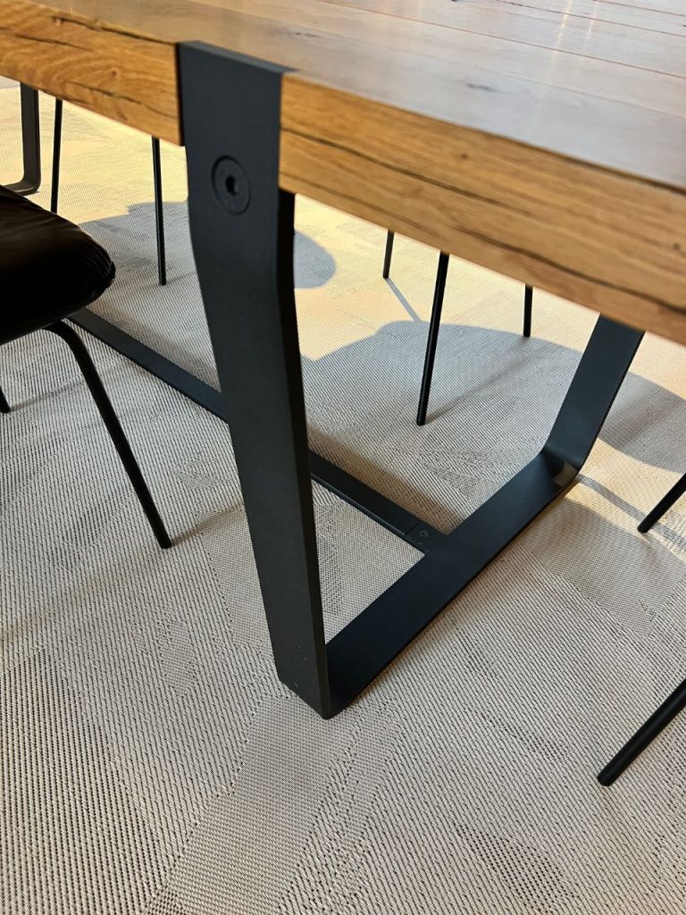 Close-up of a wooden tabletop with black metal legs and a crossbar on a light carpeted floor.