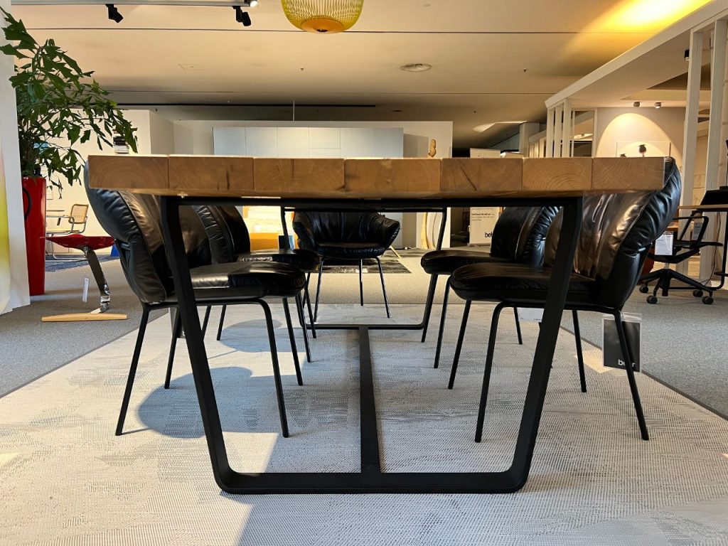 Modern conference table with a wooden top and black metal legs surrounded by black leather chairs in a bright office showroom setting.