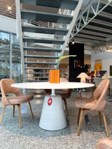 Round white table on a gray conical pedestal in a showroom, with an orange striped vase and a single green stem, surrounded by pink upholstered chairs.