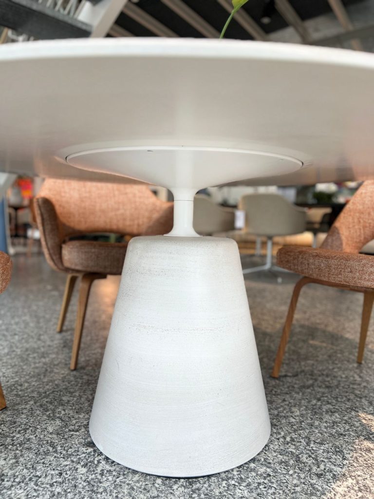 White round table with a conical pedestal base in a modern lounge area, surrounded by textured chairs on a speckled floor.
