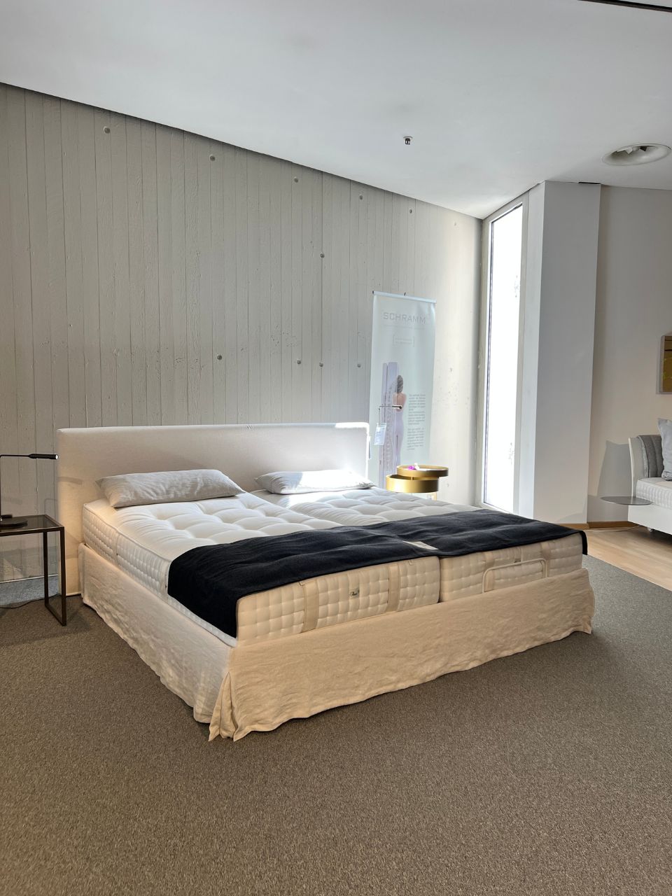 Modern showroom bedroom with a large white upholstered bed, two pillows, and a black throw blanket on white bedding. A side table and window are in view.