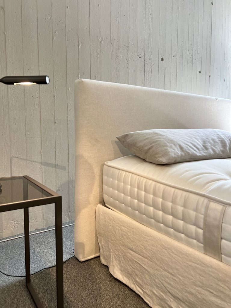 White upholstered bed with a padded headboard, gray pillow, and white quilt beside a glass-topped nightstand and a black wall lamp in a minimalist bedroom corner.