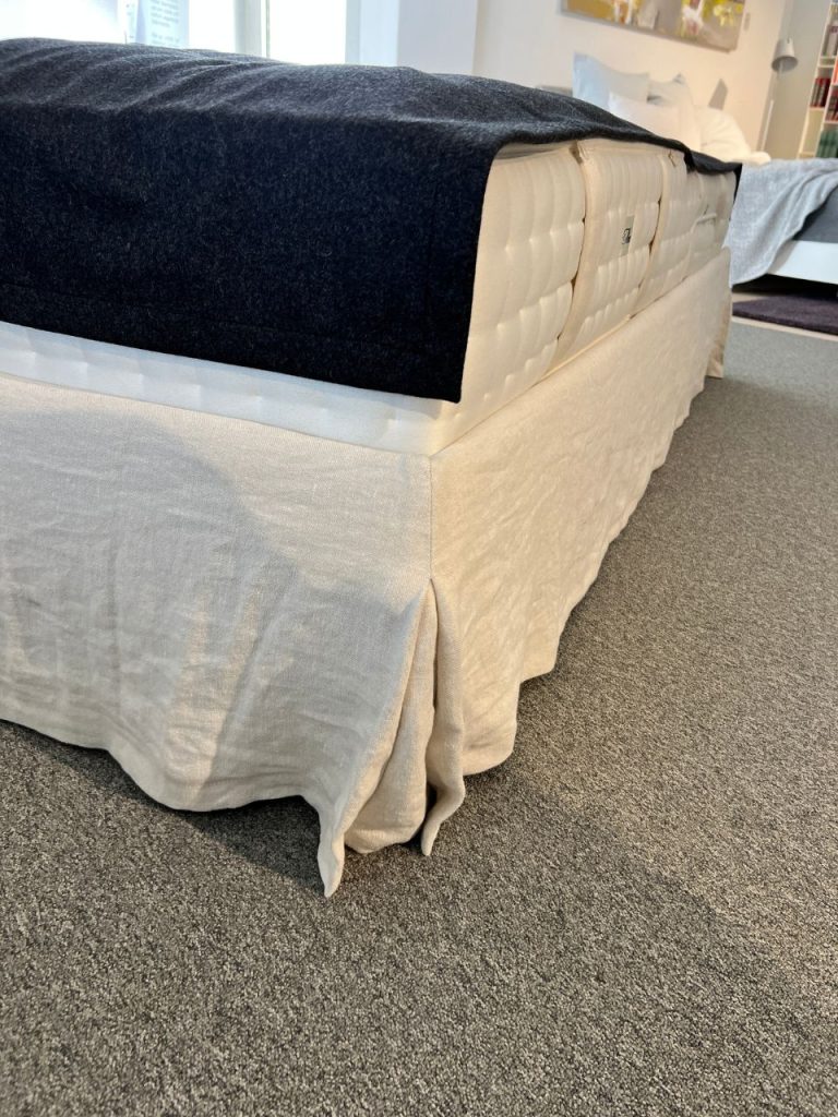 Low-angle view of a bed with a beige bed skirt and a dark blanket over a tufted white mattress on a gray carpeted floor.