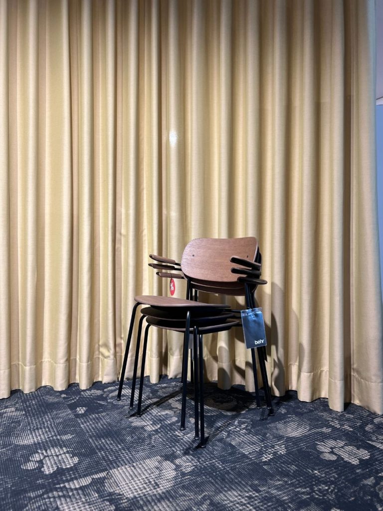 Stack of wooden chairs with black metal legs leaned together against beige curtains in a room, one chair with a blue Behr tag hanging from the back.