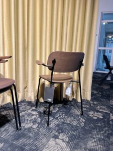 Brown wooden chair with black metal legs in front of beige curtains; tag hanging from seat reads 'behr' (furniture showroom).