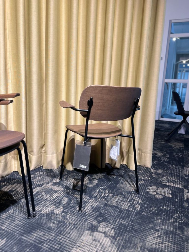 Brown wooden chair with black metal legs in front of beige curtains; tag hanging from seat reads 'behr' (furniture showroom).