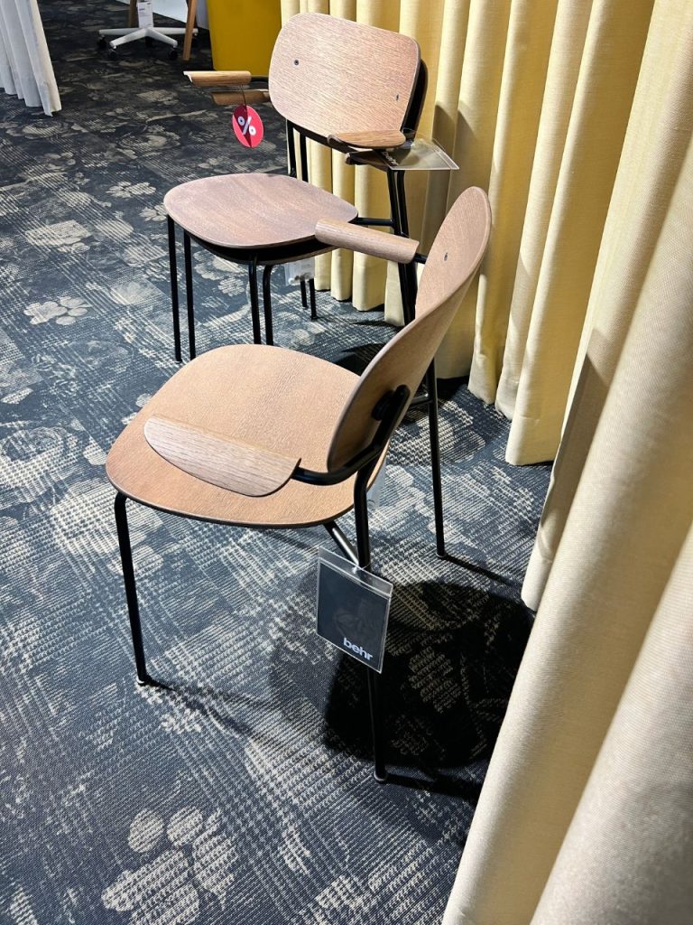 Wooden school chair with an attached writing desk, shown with another chair in a showroom with beige curtains in the background.