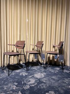 Three wooden chairs with attached desk arms, lined up in front of a beige curtain; price tags hang from the seats.