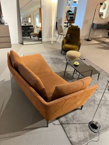 Showroom living room display: orange-brown sofa, olive armchair, and a small black coffee table on a gray rug.