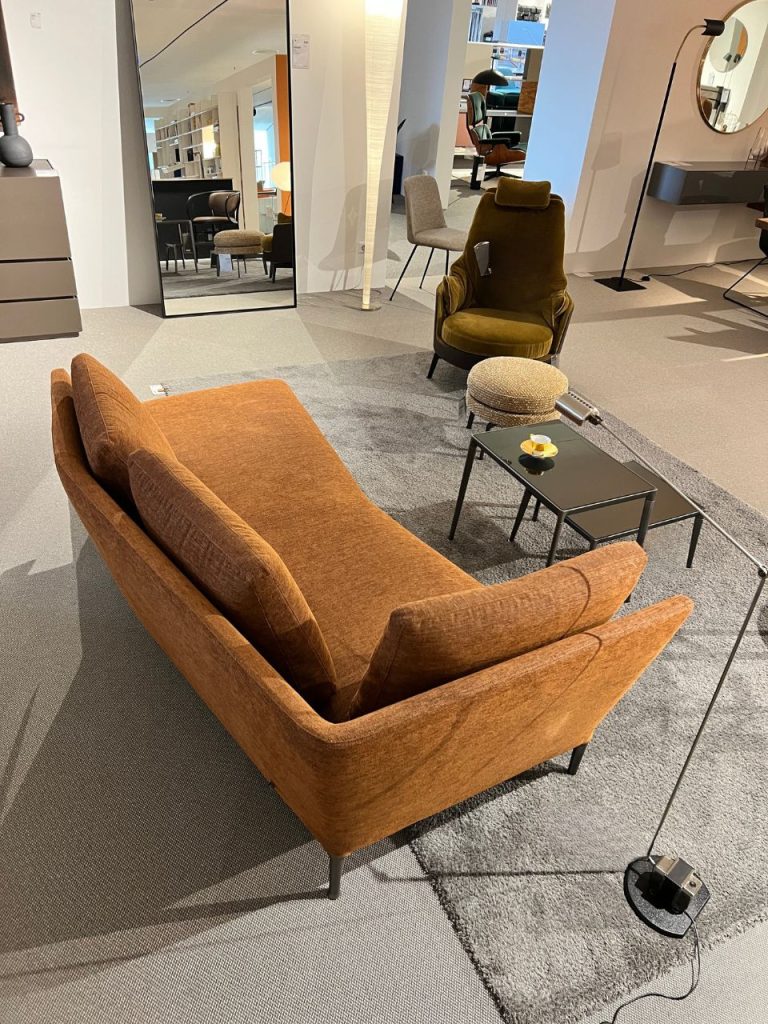 Showroom living room display: orange-brown sofa, olive armchair, and a small black coffee table on a gray rug.