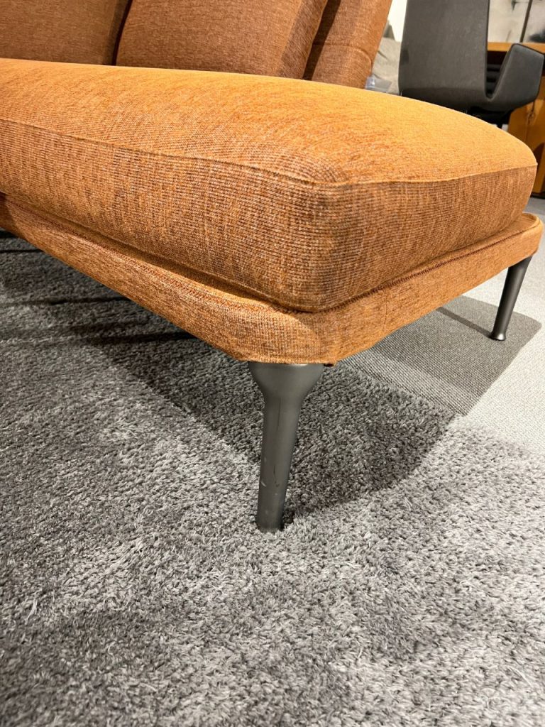 Close-up of an orange-brown textured upholstered chair seat with dark gray legs on a gray carpet in a showroom setting