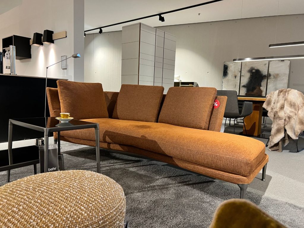Orange-brown modular sofa with a chaise on a gray rug in a modern showroom, a small glass coffee table with a cup beside it, and a fur throw draped over a chair in the background.