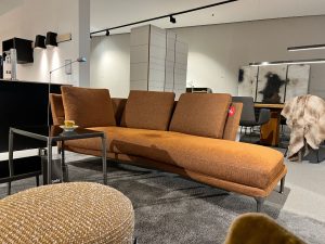 Orange-brown modular sofa with a chaise on a gray rug in a modern showroom, a small glass coffee table with a cup beside it, and a fur throw draped over a chair in the background.