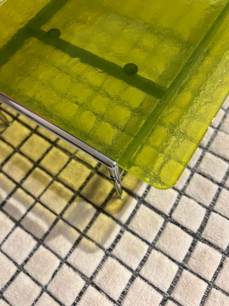 Close-up of a lime-green translucent table surface with chrome trim and leg, seen above a checkered rug.