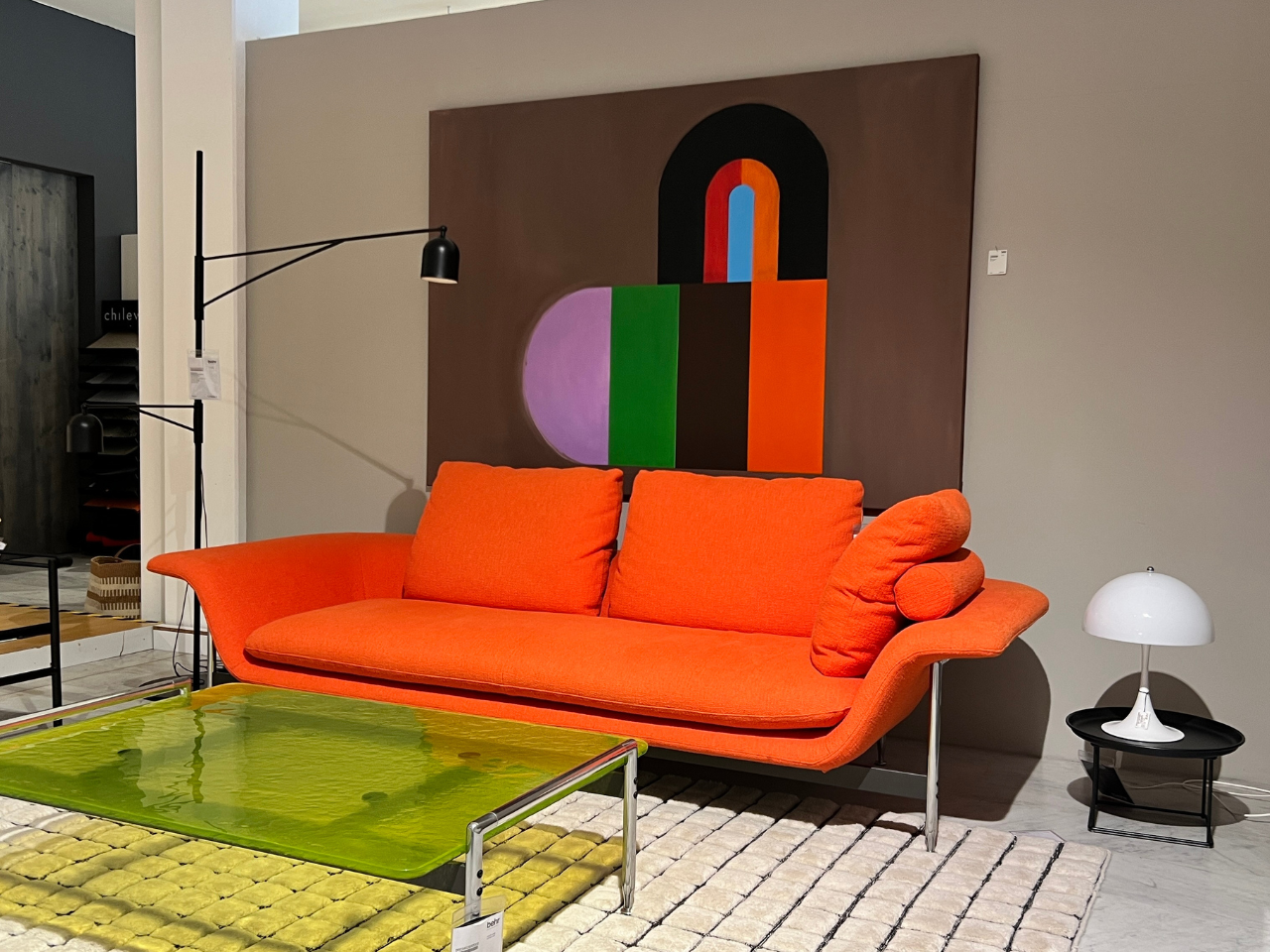 Bright orange mid-century sofa with matching cushions in a modern showroom, behind a large abstract painting with bold color blocks.