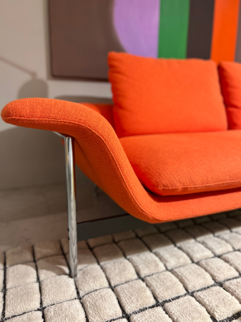 Close-up of an orange upholstered sofa arm and seat cushions on a chrome leg, set on a textured beige rug with a colorful abstract painting in the background.