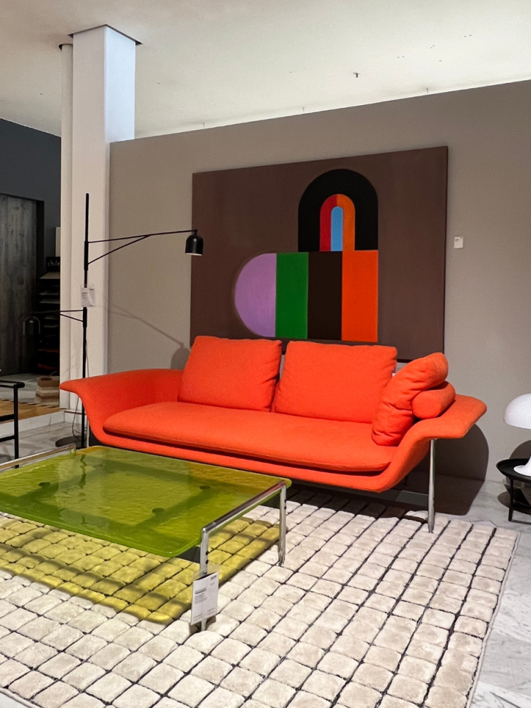 Orange modern sofa with three cushions sits on a beige rug in a showroom, next to a lime glass coffee table and a black arc floor lamp, with a colorful abstract painting on the wall behind.