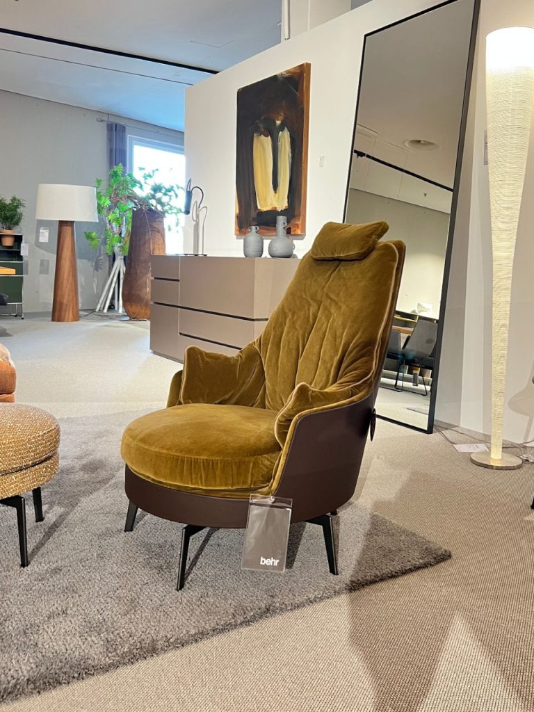 Mustard velvet lounge chair with a dark brown base on a light gray rug in a modern furniture showroom, tag hanging from the front leg.