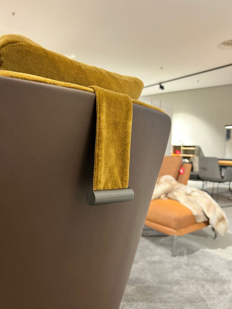 Mustard blanket draped over the back of a dark brown chair in a modern showroom. The cozy textile contrasts with the chair’s smooth leather surface, with other furniture visible in the background.