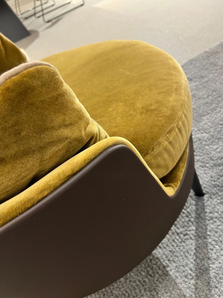 Close-up of a mustard yellow velvet chair with a curved brown armrest on a gray carpeted floor bed.