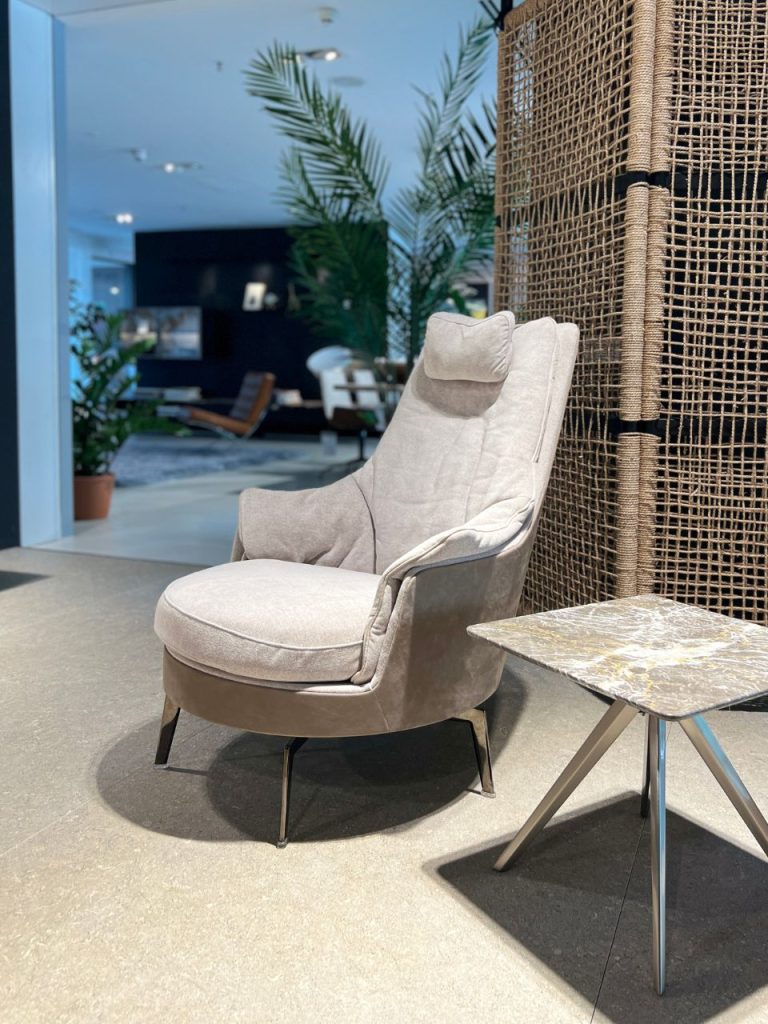 Beige upholstered lounge chair with a headrest beside a marble-topped side table in a showroom, with plants and a wicker screen in the background.