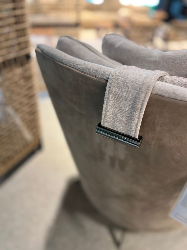 Close-up of a gray upholstered chair back with a light gray fabric strap draped over it and a shiny metal buckle/toggle at the edge.