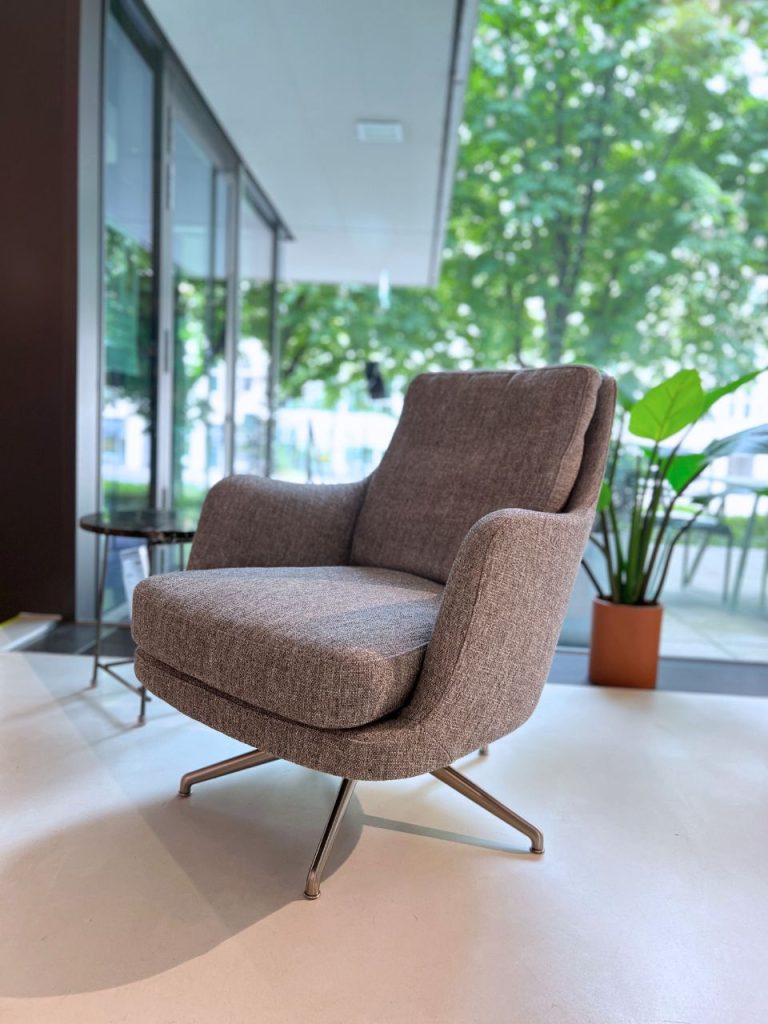 Gray upholstered swivel armchair with metal legs in a bright lobby, next to a potted plant by glass windows.