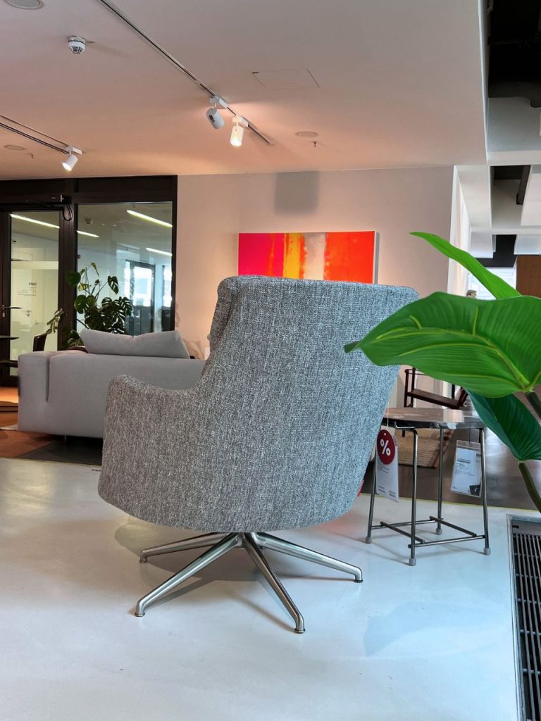 Grey upholstered swivel lounge chair with a metal four-star base in a modern showroom, with a green plant to the right and a bright abstract painting in the background.