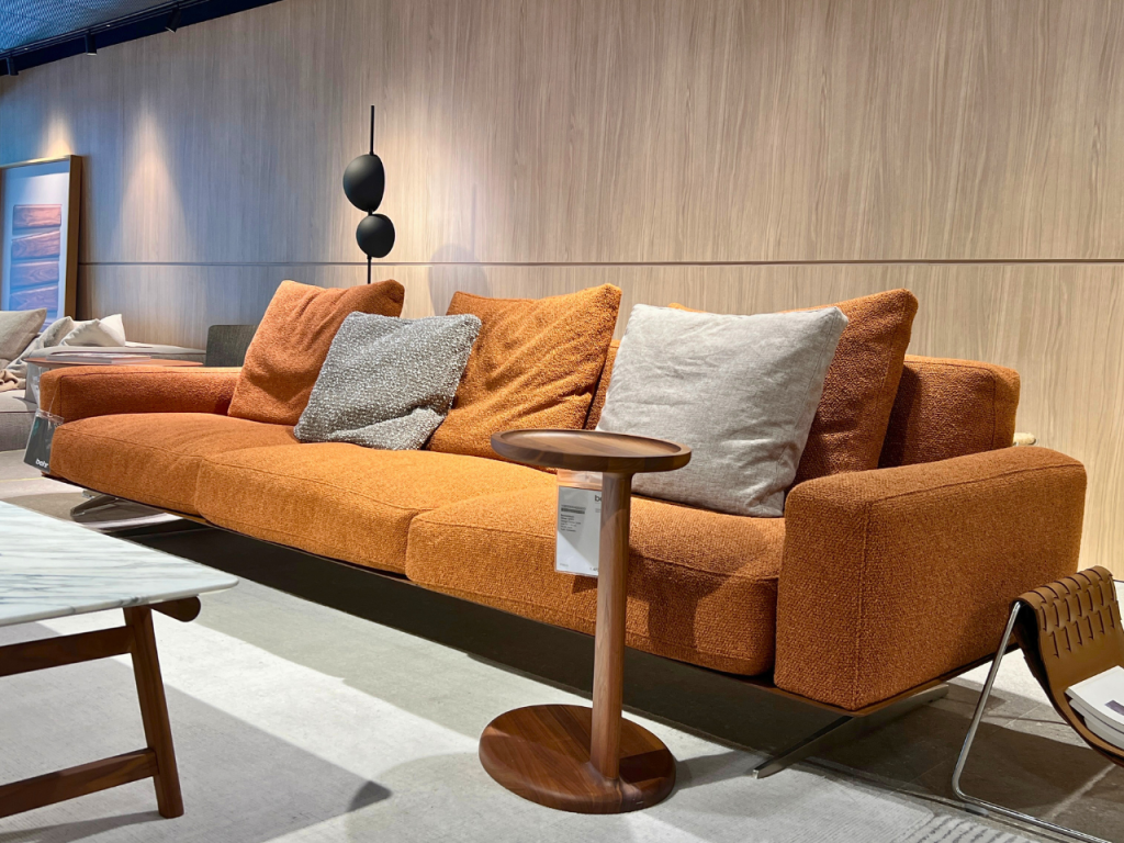 Orange fabric sofa with gray and orange cushions in a showroom, with a round wooden side table and a modern black sphere lamp behind it.