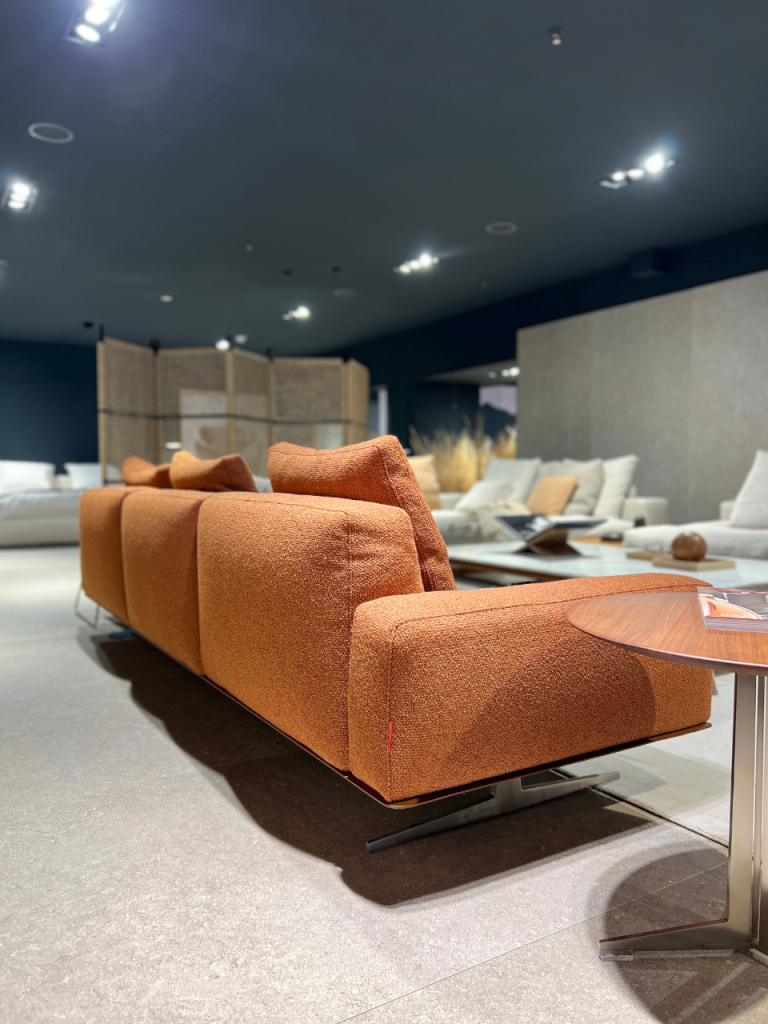 Long orange fabric sofa in a modern showroom with a round wooden table nearby.