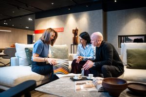 Three people on a light sofa in a showroom, a woman presenting fabric swatches to a couple as they discuss them.
