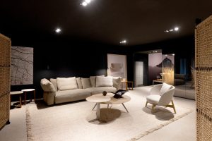 Modern living room showroom with a beige/gray sectional sofa, white chair, and round coffee tables on a light rug. Note: concise, informative description of the scene.