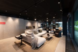 Open-concept showroom lounge with a white sectional sofa, gray cushions, and a neutral rug beneath modern coffee tables under track lighting.
