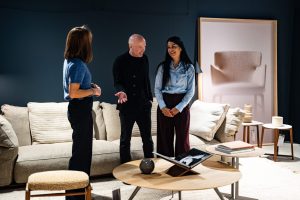 Three adults in a showroom lounge talk beside a beige sofa and round coffee tables.
