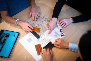 Three people collaborate around a wooden table, comparing fabric swatches, a carpet sample, and a ruler over blueprints with a tablet displaying a design site nearby.