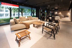 Modern showroom seating area with a beige sectional sofa, woven chairs, and a wooden coffee table by large windows overlooking trees in a furniture store.