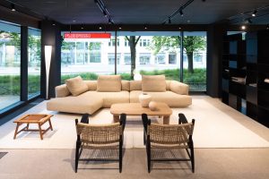 Modern living room showroom with a beige sectional sofa, woven chairs, and a wood coffee table by large floor-to-ceiling windows.