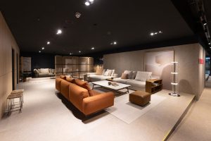 Modern furniture showroom with an orange sectional sofa and white sofas around a rectangular coffee table, warm lighting overhead.